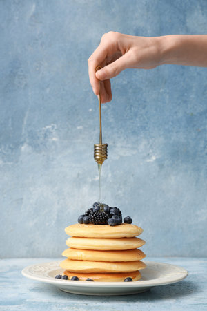 Woman pouring honey on sweet pancakes with fresh blueberries and blackberries against light blue...