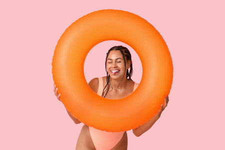 Beautiful young African-American woman in stylish swimsuit with inflatable ring showing tongue on...