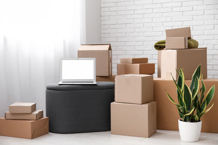 Blank laptop on pouf with moving boxes in light room