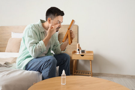 Young man with mirror putting in contact lenses at homeの写真素材