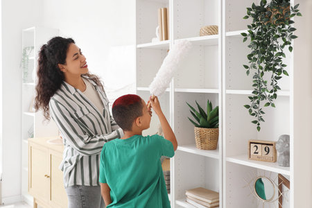 Little African-American boy with his mother cleaning shelf at home