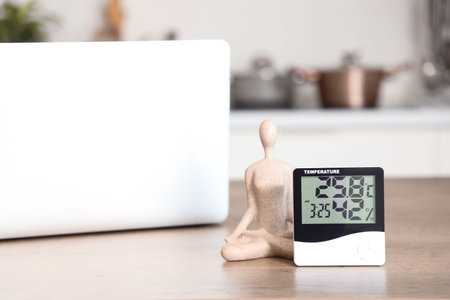 Digital hygrometer with statuette and laptop on table in kitchen, closeup