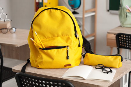 School desk with eyeglasses, backpack and notebook in light classroomの写真素材