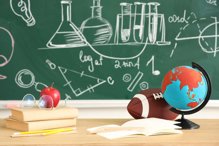 School desk with apple, books, globe and rugby ball in light classroom
