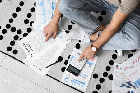 Young man in debt using calculator on floor at home, top view