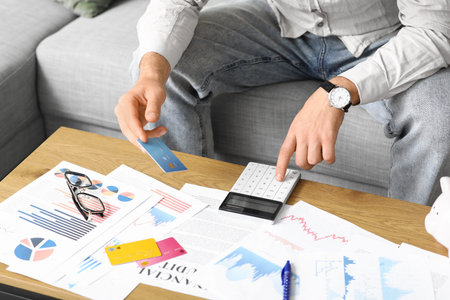 Young man in debt with credit card using calculator at home, closeup