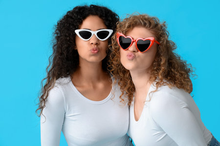 Beautiful young women in sunglasses blowing kiss on blue background