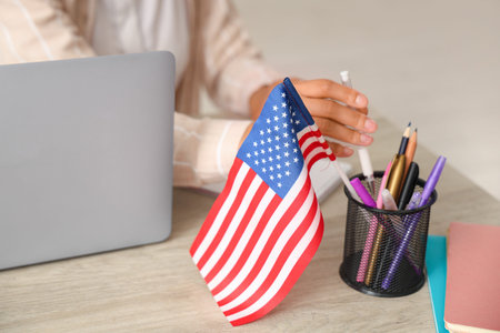 Beautiful young African-American businesswoman with USA flag and laptop sitting at table in office,...