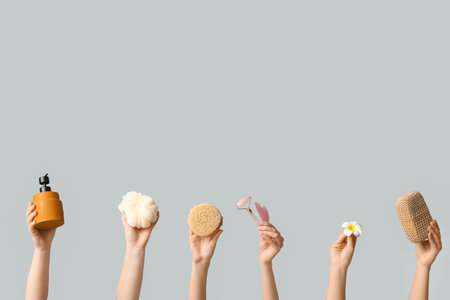 Female hands with bath sponges, massage body brushes, facial massage tools, flower and bottle of cosmetic product on gray backgroundの写真素材