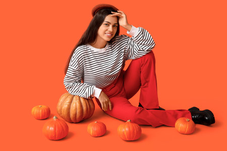 Stylish young woman with fresh pumpkins sitting on orange backgroundの写真素材