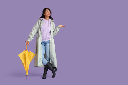 Happy young African-American woman in raincoat with umbrella on lilac backgroundの写真素材
