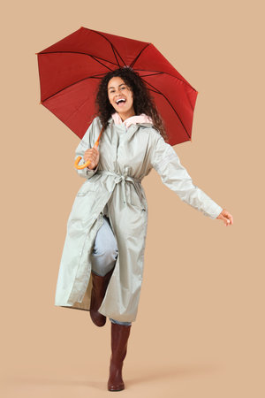 Happy young African-American woman in raincoat with umbrella on beige backgroundの写真素材
