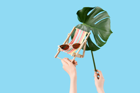 Female hands with sunglasses, mini deck chair and monstera leaf on blue background. Travel conceptの写真素材