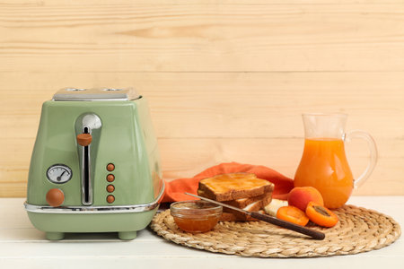 Green toaster with bread slices, apricots, jam and jug of juice on white wooden tableの写真素材