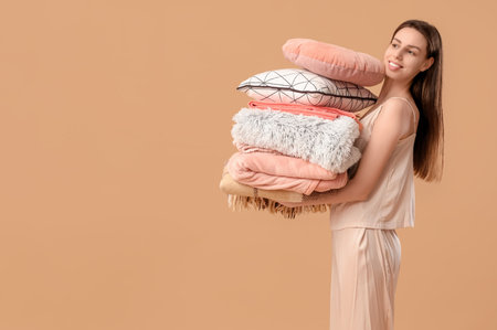 Beautiful young happy woman with stack of different blankets and pillows on beige backgroundの写真素材
