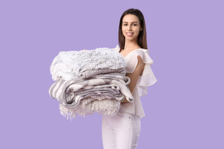 Beautiful young happy woman with stack of different blankets on purple background