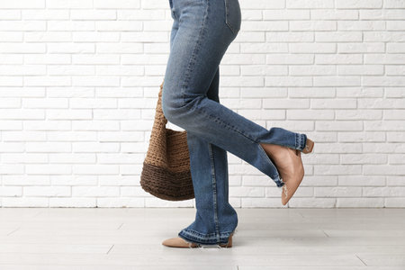 Stylish young woman in jeans and shoes with bag near white brick wall