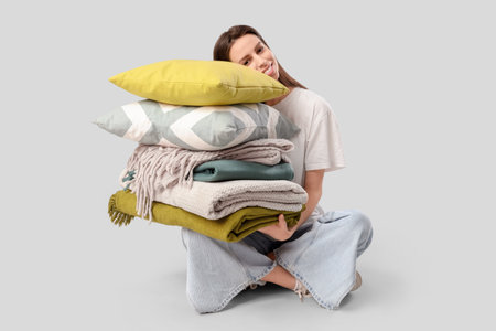 Beautiful young happy woman with stack of different blankets and pillows sitting on gray backgroundの写真素材