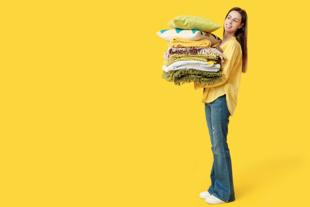 Beautiful young happy woman with stack of different blankets and pillows on yellow backgroundの写真素材