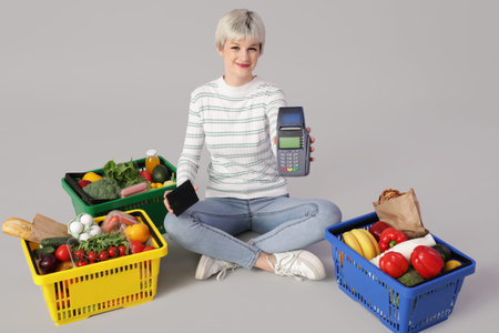 Young woman with payment terminal, wallet and full shopping baskets on light background