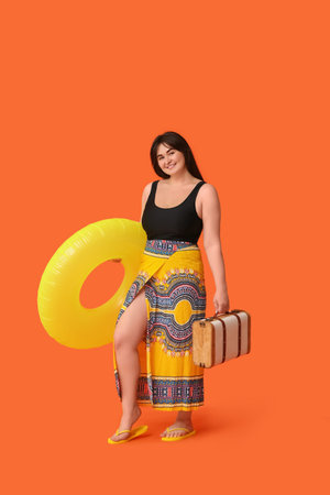 Beautiful young woman with inflatable ring and suitcase on orange background
