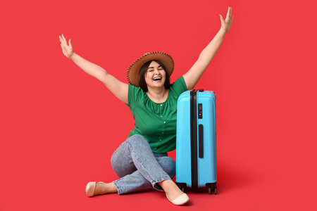 Happy young woman with suitcase sitting on red backgroundの写真素材