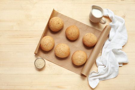Baking paper of fresh buns with sesame seeds and jug of milk on wooden background
