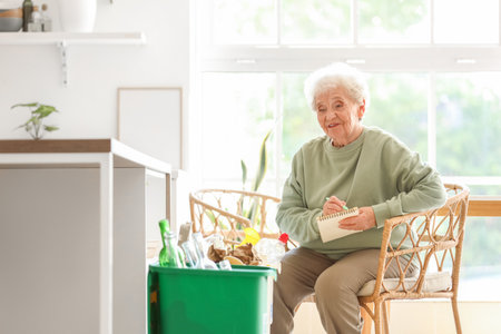 Senior woman with garbage bins writing on notebook in kitchen. Waste sorting conceptの写真素材