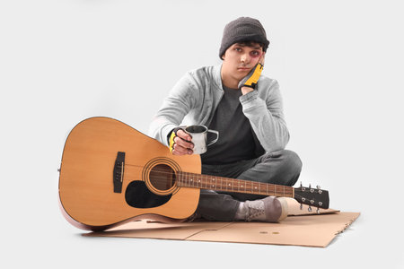 Young homeless man with cup and guitar begging for money on light backgroundの写真素材