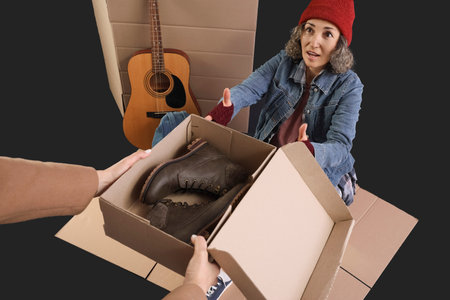 Mature homeless woman receiving donation shoes on dark background