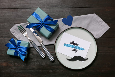 Festive decorated table setting with gift box and greeting card on black wooden background. Father...