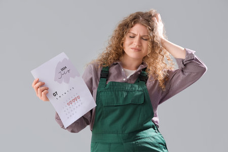 Female worker with menstrual calendar suffering from headache on gray background