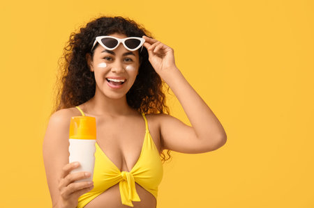 Beautiful young happy African-American woman with bottle of sunscreen cream on yellow background
