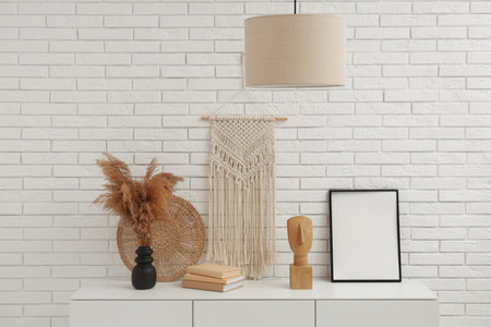 Dried flowers in vase, books and empty frame on cabinet near white brick wall