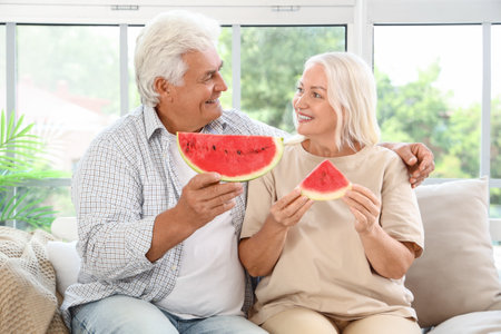 Mature couple with slices of watermelon sitting on sofa at homeの写真素材