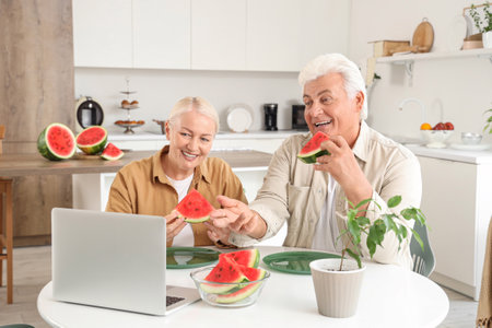 Mature couple with slices of fresh watermelon video chatting at table in kitchenの写真素材
