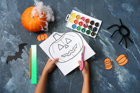 Child with supplies cutting paper pumpkin while making Halloween decorations on grunge blue backgroundの写真素材