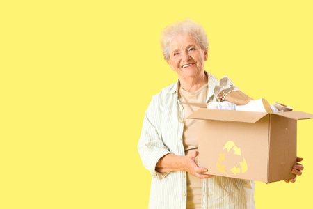 Senior woman with box of garbage on yellow background. Waste sorting concept
