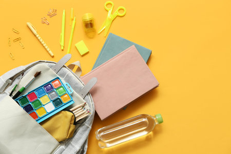 Modern school backpack with different stationery supplies and bottle of water on yellow background