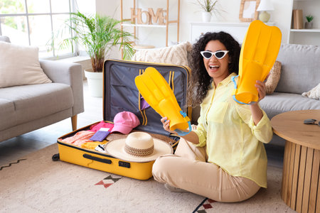 Young African-American woman unpacking suitcase with traveling accessories and flippers in hotel roomの写真素材