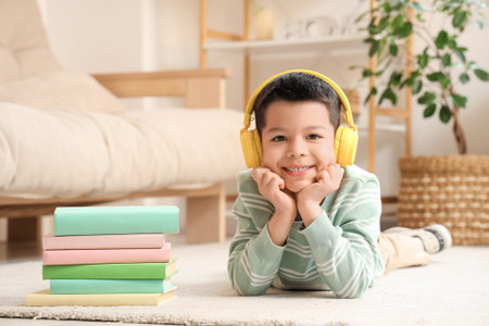 Cute little Asian boy in headphones listening to audiobook with books while lying on carpet in...