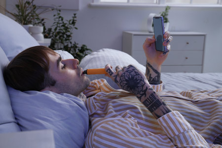 Young man in pajamas smoking disposable electronic cigarette and using mobile phone on bed at homeの写真素材