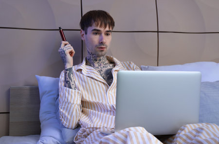 Young man in pajamas with disposable electronic cigarette using laptop on bed at homeの写真素材