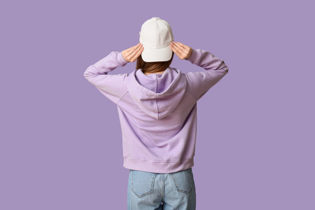 Young woman in stylish white cap on lilac background, back view