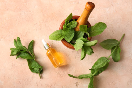 Bottle of essential mint oil, mortar, pestle and green leaves on pink grunge background