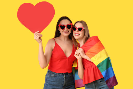 Beautiful young lesbian couple with LGBT flag and paper heart on yellow backgroundの写真素材