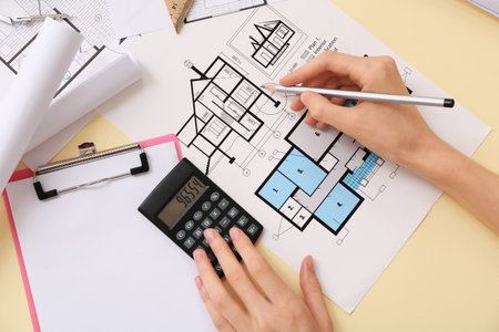 Female architect with house plan using calculator on yellow background, top view