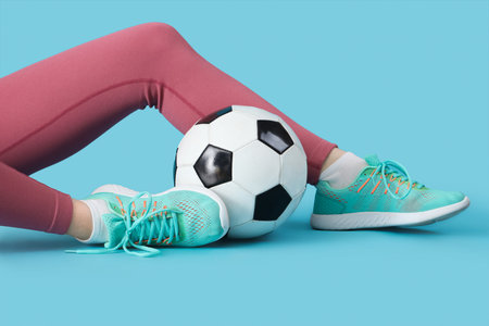 Female legs in stylish sneakers with soccer ball on blue background