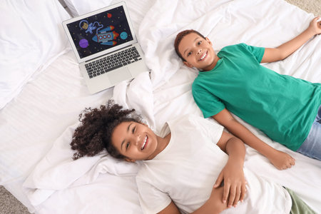 Cute African-American children watching cartoons on laptop in bedroom at home, top view