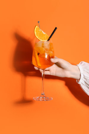 Female hand with glass of Aperol spritz cocktail on orange backgroundの写真素材